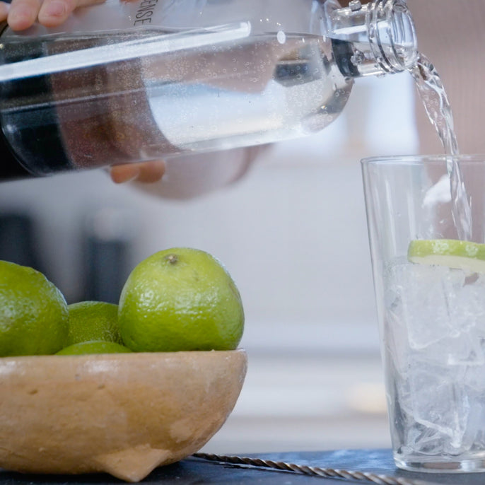 A hand pours sparkling water from a Soda Sense bottle into a glass filled with ice and lime slices. In the foreground, a wooden bowl holds several fresh limes.