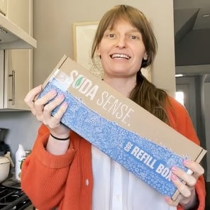 A customer smiling while holding a Soda Sense CO2 canister refill box, ready to be mailed for exchange.