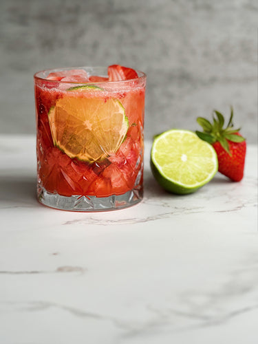 A strawberry mocktail in a clear, textured glass with vibrant red liquid, ice, strawberry slices, and a lime round. To the side, there's a half lime and a strawberry on a white marble countertop.